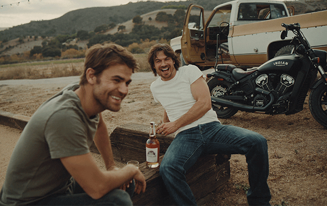 韦斯利和萨默海尔德坐在摩托车旁，手里拿着一瓶兄弟的邦德波旁威士忌